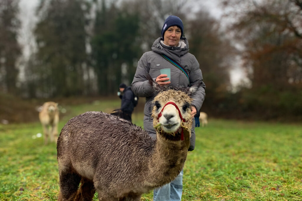 Frau mit einem Becher blickt in die Kamera, davor steht ein Alpaka